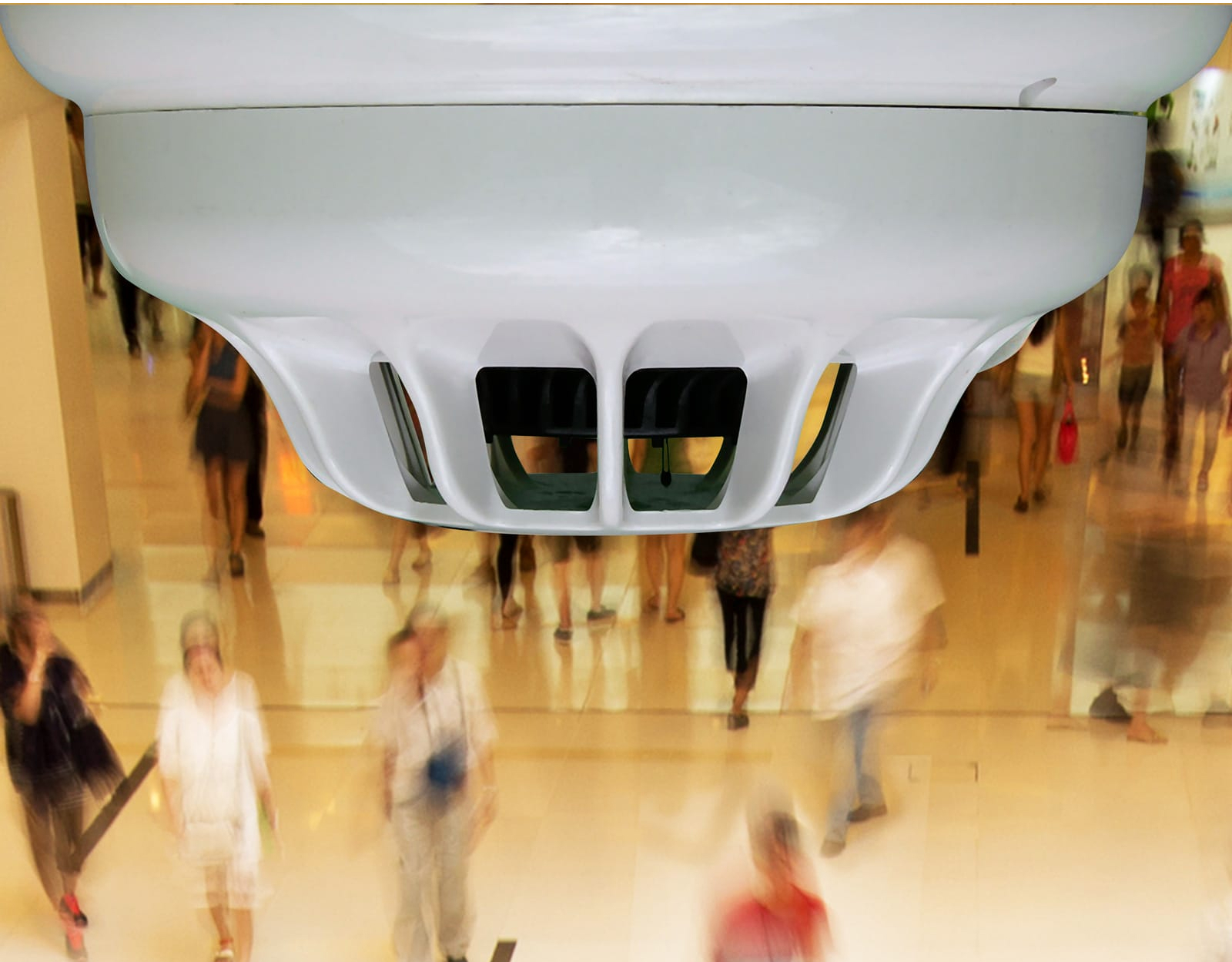 Close-up of a fire detector mounted on the ceiling, with people walking below in the background